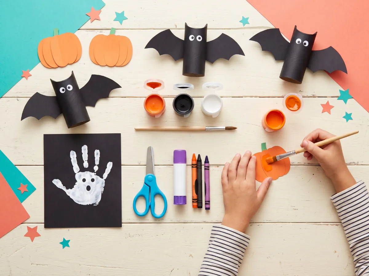 Kids Halloween craft flat-lay with paper pumpkins, toilet-roll bats and small hands painting